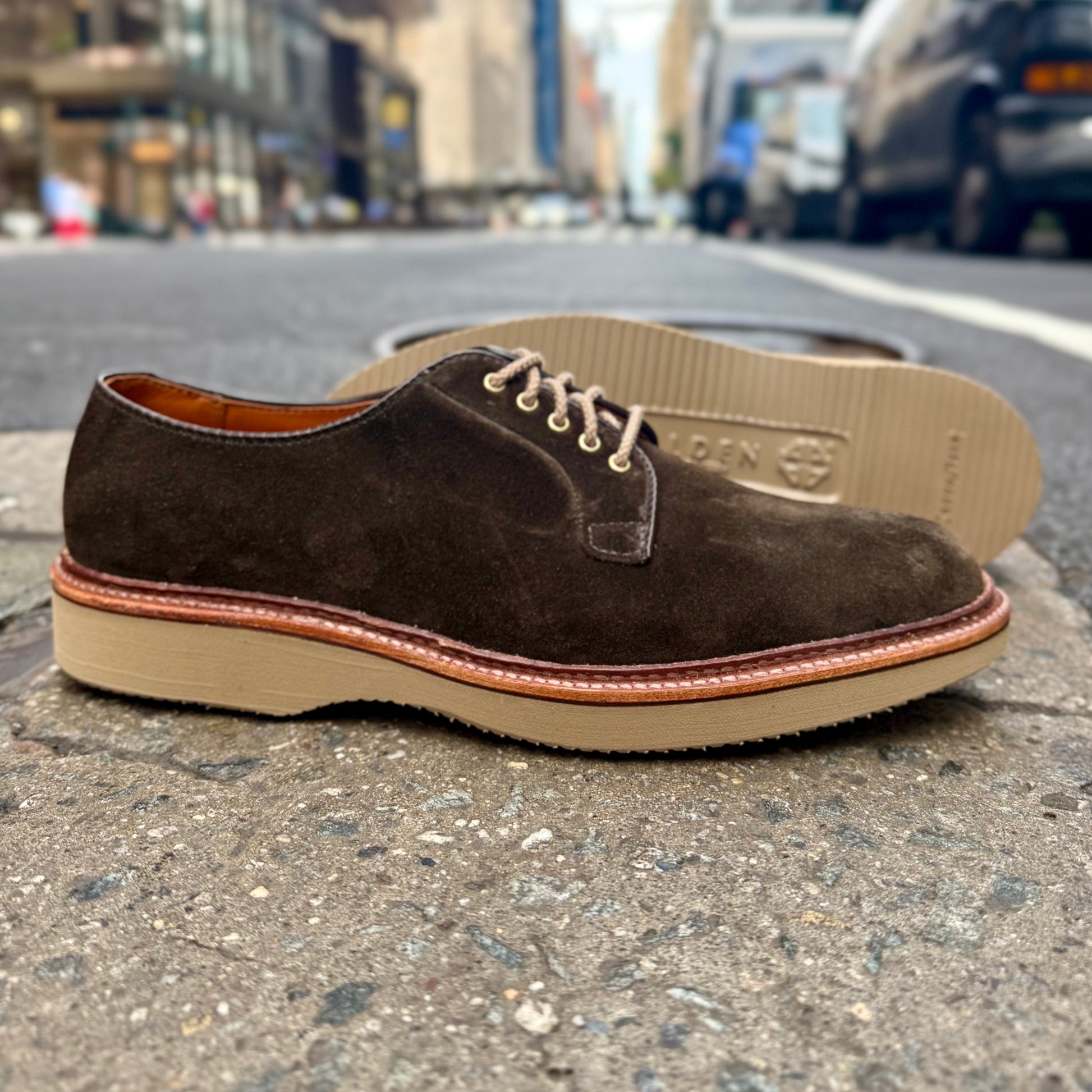 The ALDEN SHOES D5426 - Loden Suede Plain Toe Blucher, featuring beige laces and a light sole, is shown on a city sidewalk with blurred buildings and cars in the background.