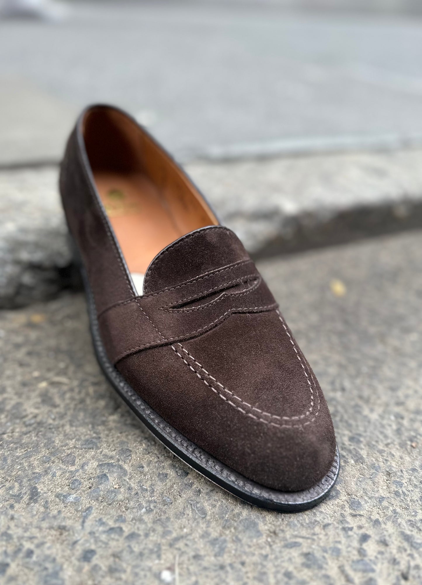The ALDEN SHOES G9201 - Dark Brown Suede Full Strap Loafer, featuring visible stitching and crafted on the Aberdeen last, rests on a rough gray stone surface near a curb.