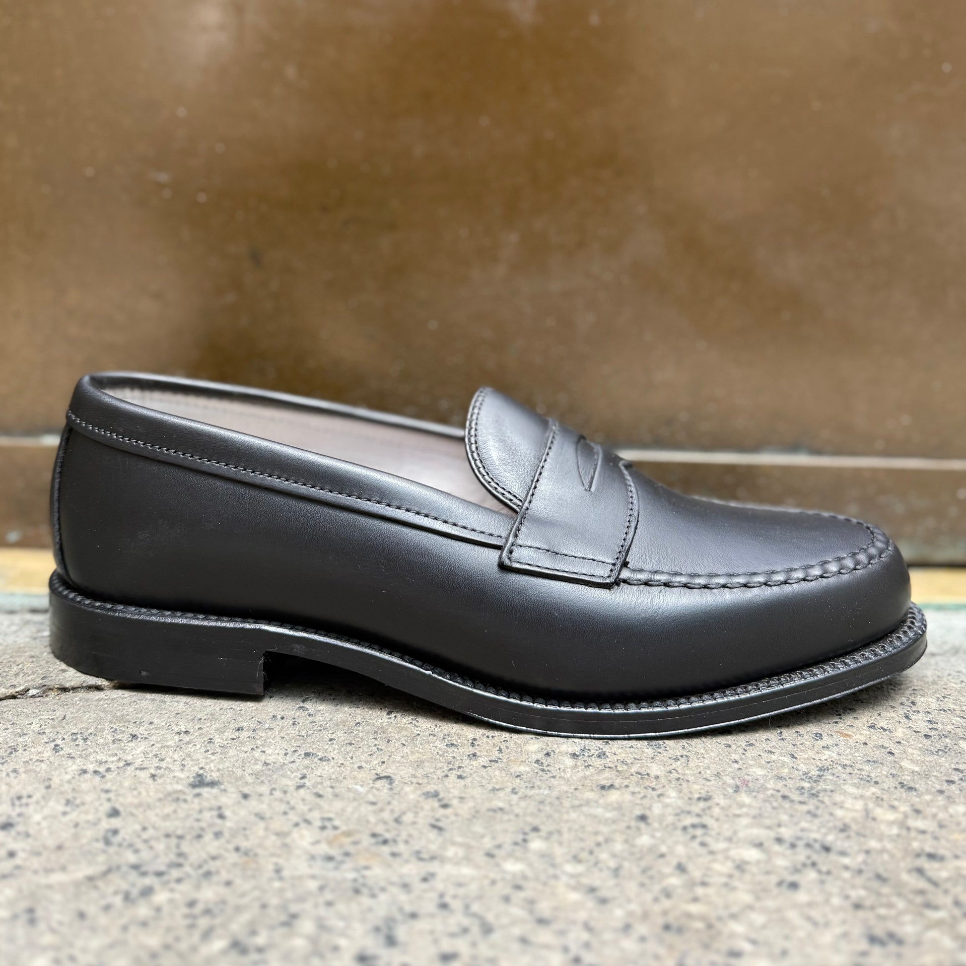 The ALDEN SHOES 9817 - LHS in Black Trapper Calf penny loafer, hand sewn with precision, is displayed on a concrete surface against a blurred brown background.