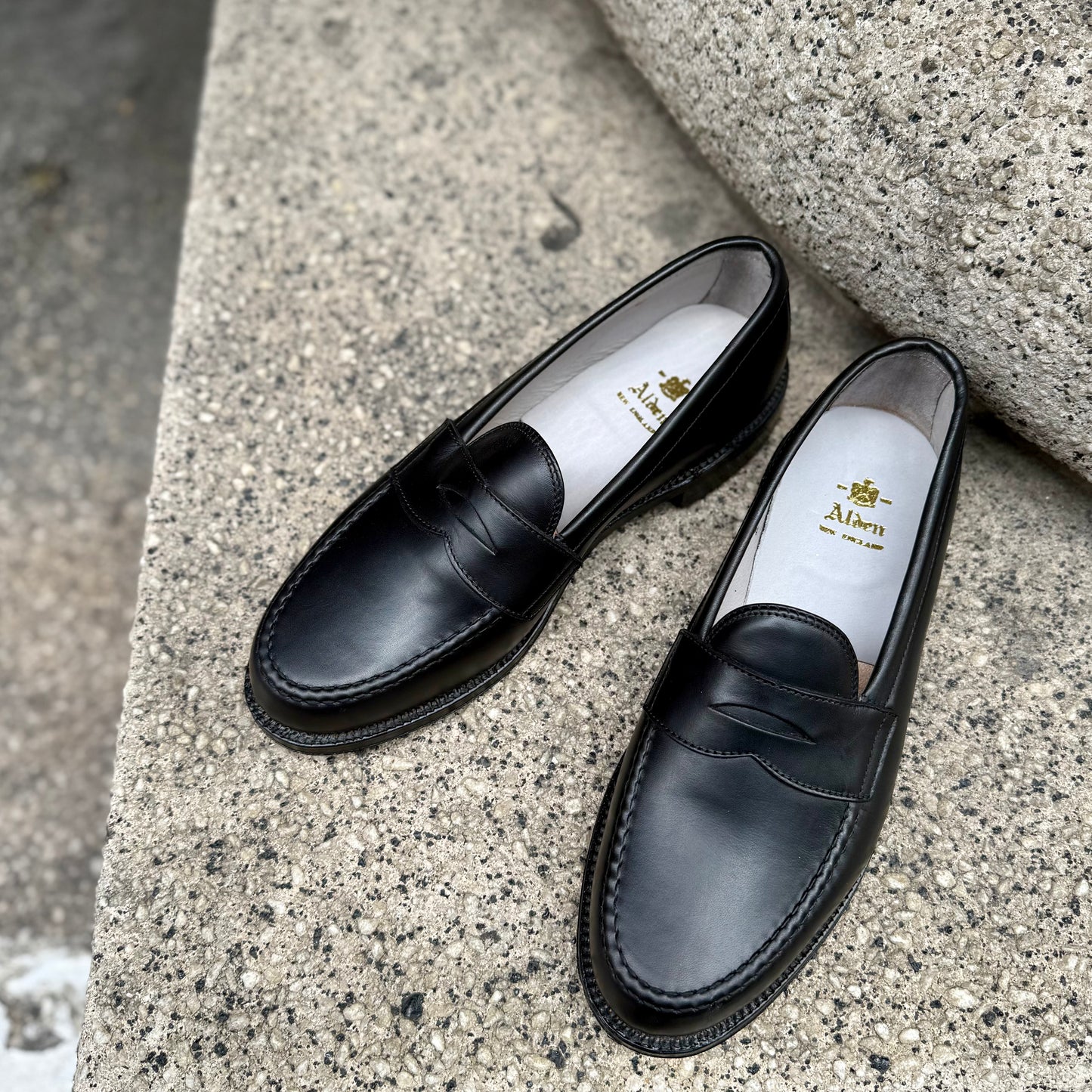 The ALDEN SHOES 9817 - LHS in Black Trapper Calf penny loafers, hand sewn with white insoles and gold "Alan" logo, are displayed on a speckled concrete surface.