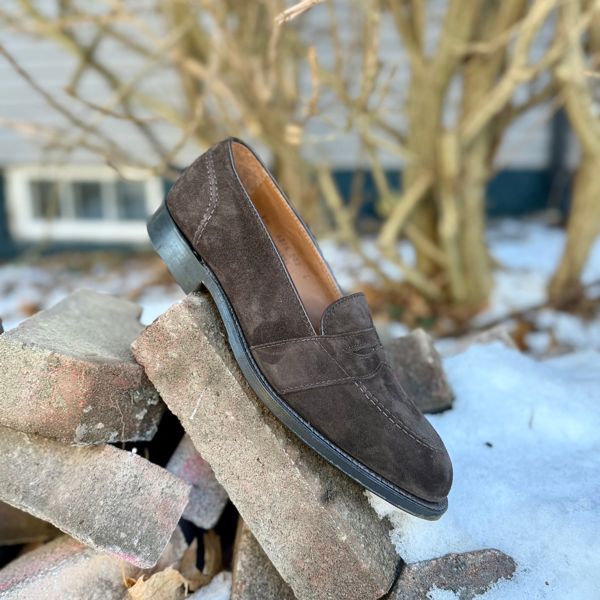 The ALDEN SHOES G9201 - Dark Brown Suede Full Strap Loafer, built on the Aberdeen last, sits atop bricks outdoors with snow and bare branches in the background.