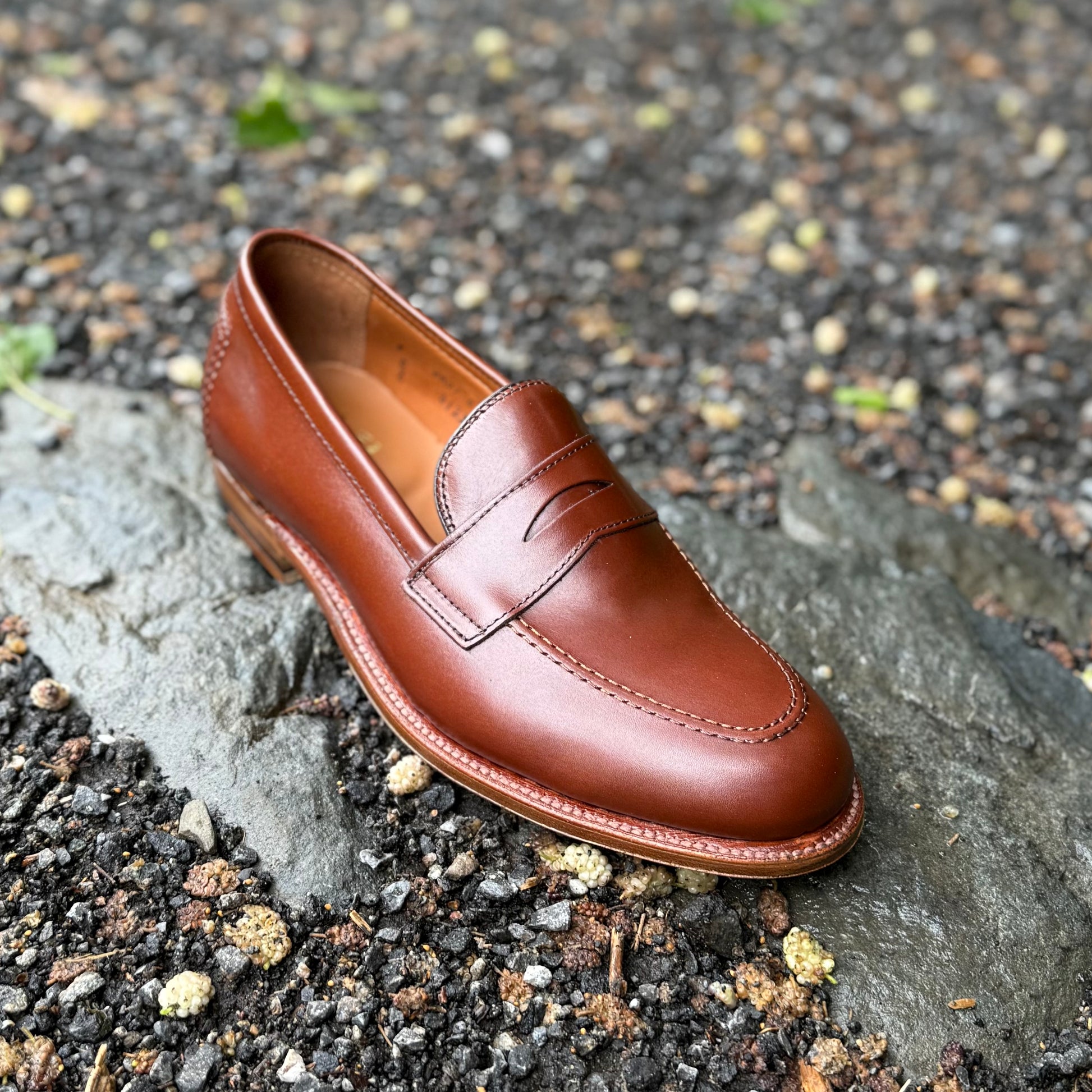 An Alden Madison D1235 - Madison Penny in Madison Brown Calfskin rests on a flat rock amid gravel and leaves, showcasing detailed stitching, a penny strap upper, and a sturdy leather sole.