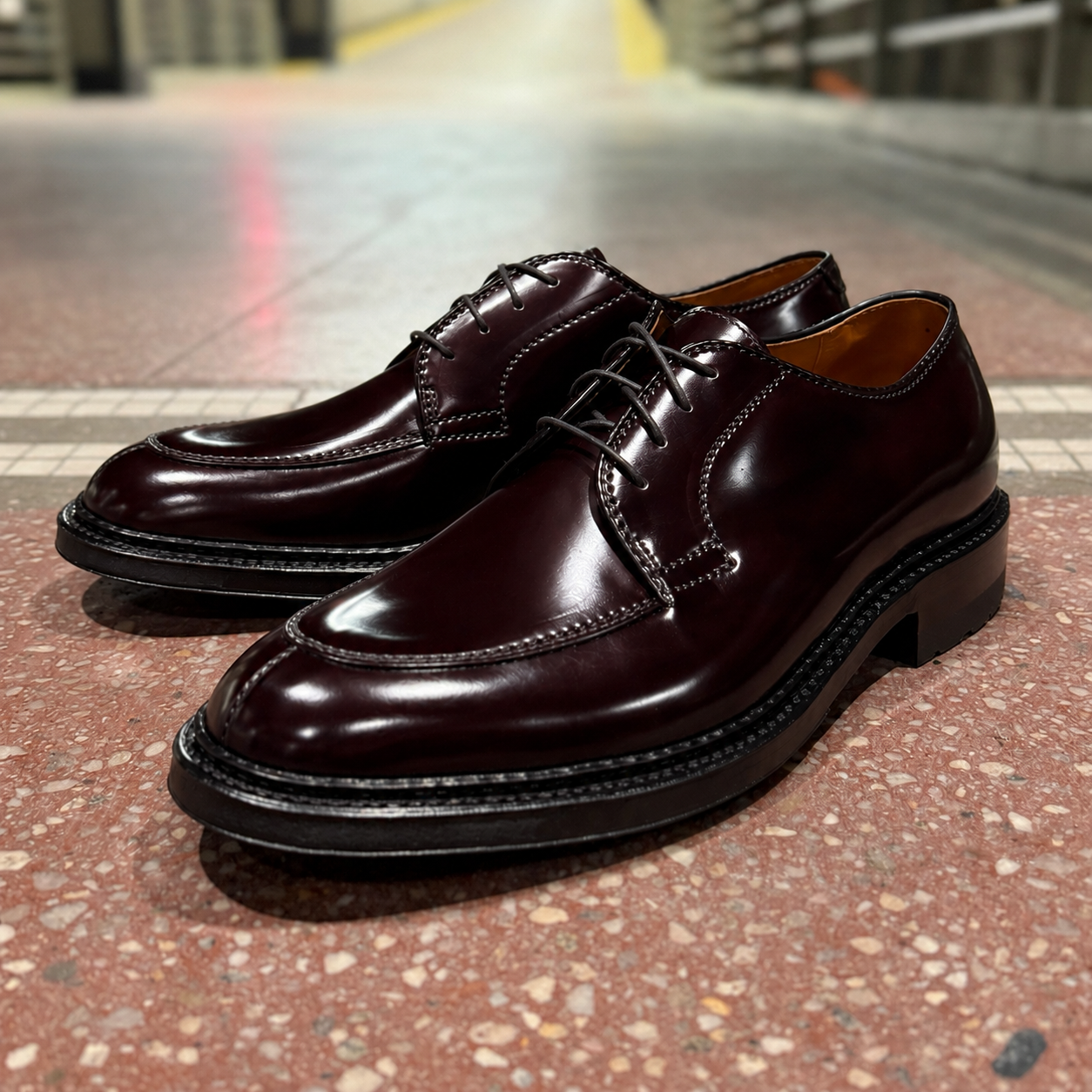 A pair of ALDEN SHOES D6632C - Borough Blucher in Color 8 Shell Cordovan leather, featuring black soles, is displayed on a speckled tile floor with a softly blurred indoor background.