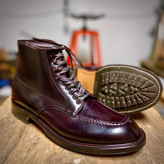 The 40508HC - Color 8 Shell Indy Boots by Alden Madison, crafted from polished dark brown leather, are displayed on wood. One boot stands upright with detailed stitching and laces; the other lies sideways exposing the textured Commando Sole, against a blurred background.