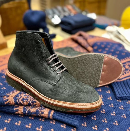 A single Alden Madison 86065H Earth Reverse Plain Toe Boot with purple laces is displayed atop a folded geometric sweater. The black suede boot features a crepe sole with gray and brown hues. Blurred clothing items appear in the background.