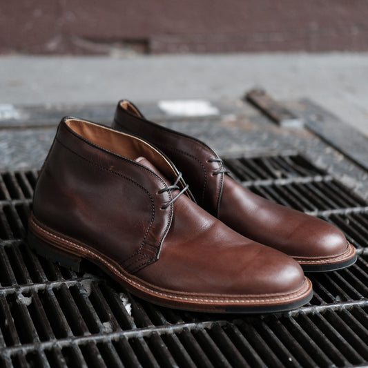 A pair of Alden Madisons 13781C Chukka Boots in Brown Chromexcel with a polished finish and visible stitching rest on a metal grate outdoors. Featuring laces and durable Commando soles, the boots are emphasized against the blurred background.