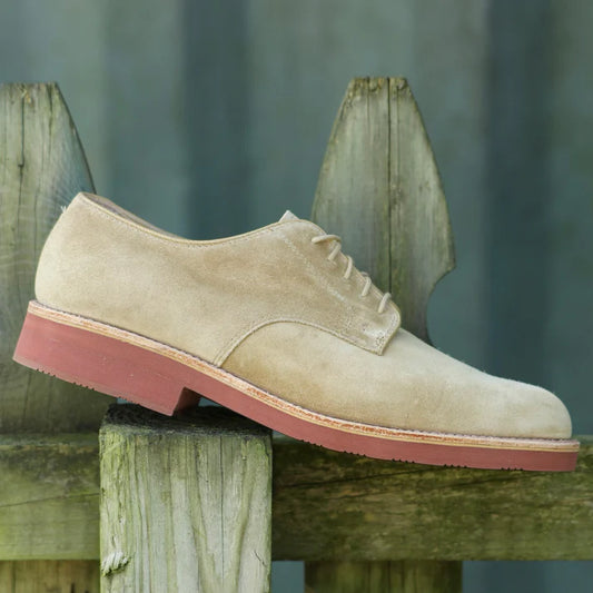 A single Alden Madison 96803 - Dirty Buck in Tan Suede shoe with a red rubber sole is resting on a wooden fence. The blurred background highlights its lightweight, classic design.