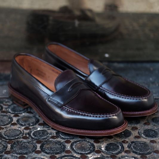 A pair of D6207 - Antique LHS in Color 8 Shell penny loafers from Alden Madison sits on a textured black rubber mat, highlighting their sleek design, leather sole, and light tan interior. The shoes feature intricate antique edging and detailed stitching, reflecting beautifully off the surface behind them.
