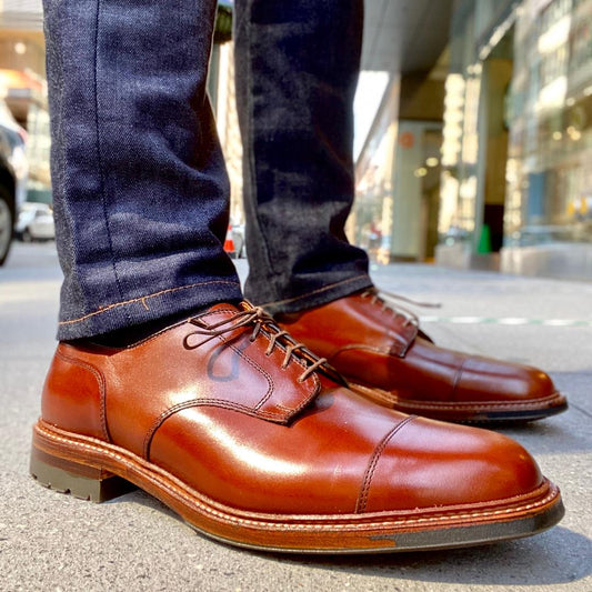 A close-up shows someone wearing Alden Madisons D9539C Ten to One DeRosa Bluchers in Madison Brown Calfskin with dark jeans. The city sidewalk setting highlights the polished, lace-up style of these Cap Toe Bluchers against the backdrop of urban buildings.