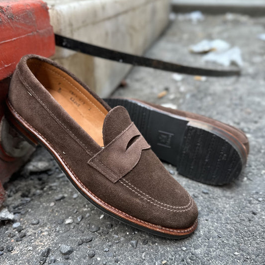 The Alden Madison 6224L Unlined Penny Mocc in Brown Suede is displayed on a textured concrete surface, with one shoe leaning against a curb to highlight its rubber sole and design features, set against a softly blurred urban background.