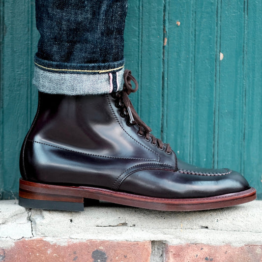 A person in dark blue cuffed jeans and Alden Madisons 40538H Indy Boot w Neoork Sole in Color 8 Shell stands on a brick ledge before a green wooden wall.