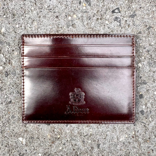 The LG 6008 - Alden Color 8 Shell Credit Card Holder by Alden Madison is a dark brown leather cardholder with three slots, embossed with Albert and a crest. It rests on a textured gray concrete surface, epitomizing refined style and craftsmanship.