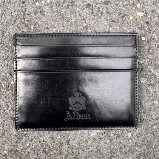 The Alden Madison LG 6009 is a black leather Shell Cordovan cardholder embossed with Alden and a crown logo, featuring six pockets for essentials, elegantly displayed on textured stone.