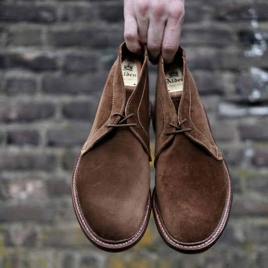 An Alden Madison hand holds a pair of 1493 - Unlined Chukka in Snuff Suede boots against a rustic brick wall.