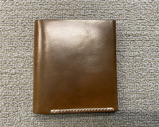 The LG 7007 - Alden Cognac Shell Cordovan Wallet by Alden Madison, with a closed brown leather design and white stitching, sits elegantly on a gray textured surface.