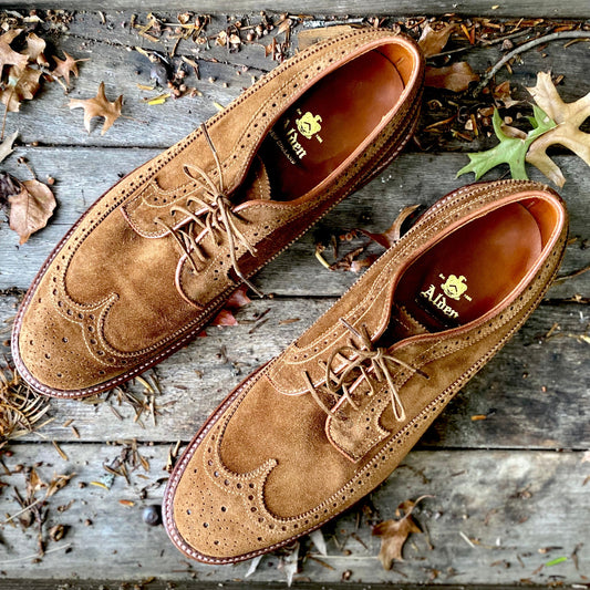 A pair of Alden Madison 9794 Long Wing Blucher in snuff suede, crafted on the Barrie last with a Waterlock leather sole, sits on a wooden surface. Detailed stitching and fallen leaves create an autumnal scene.