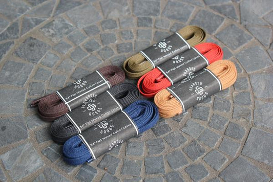 Packets of colorful shoelaces, including Alden Madisons Flat Boot Lace in 54 inch Black and Brown, are arranged on a circular stone-patterned surface. Each pair is neatly coiled and wrapped in branded packaging.