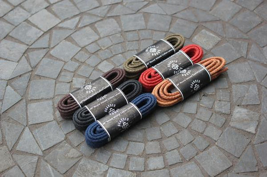 Six pairs of 48-inch Alden Madison Round Boot Laces in black, tan, olive, red, brown, and orange sit neatly coiled on a stone-tiled surface. Each pair is wrapped with a black band featuring the brand name.